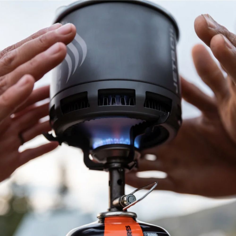 Person holding a camping stove with a visible flame, outdoors.