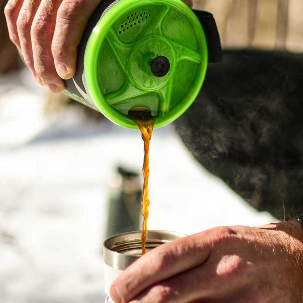 Person pouring coffee from a green and black coffee maker into a silver cup.