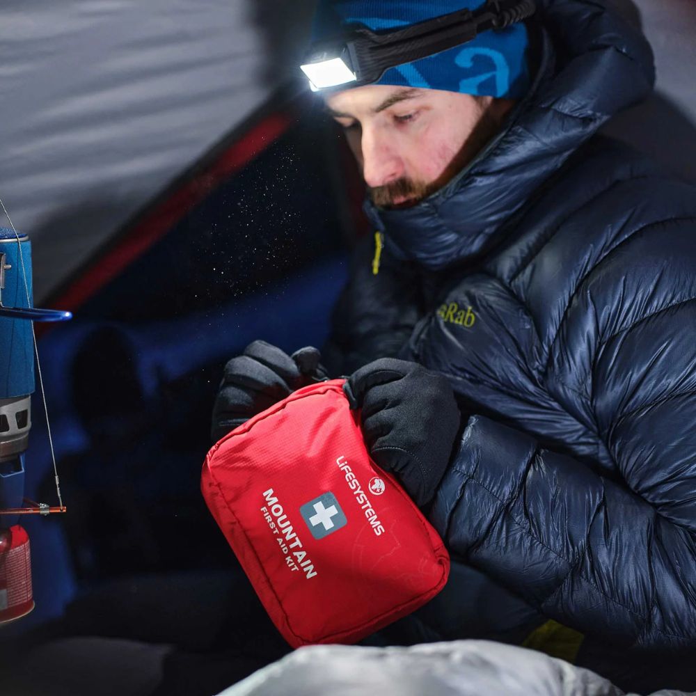 Person in winter clothing with a red Lifesystems Mountain First Aid Kit inside a tent.