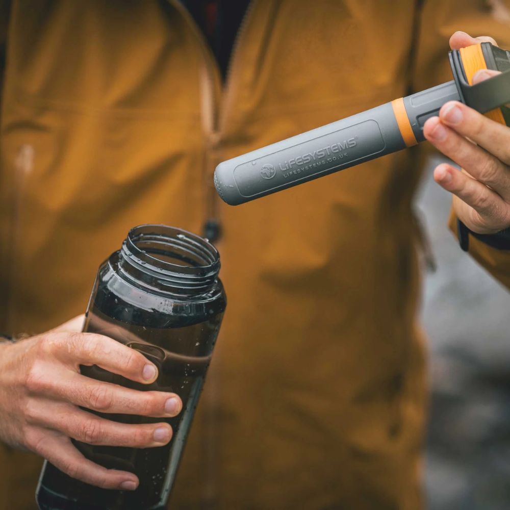 Person holding a black water bottle and a LifeStraw filter in a blurred outdoor setting