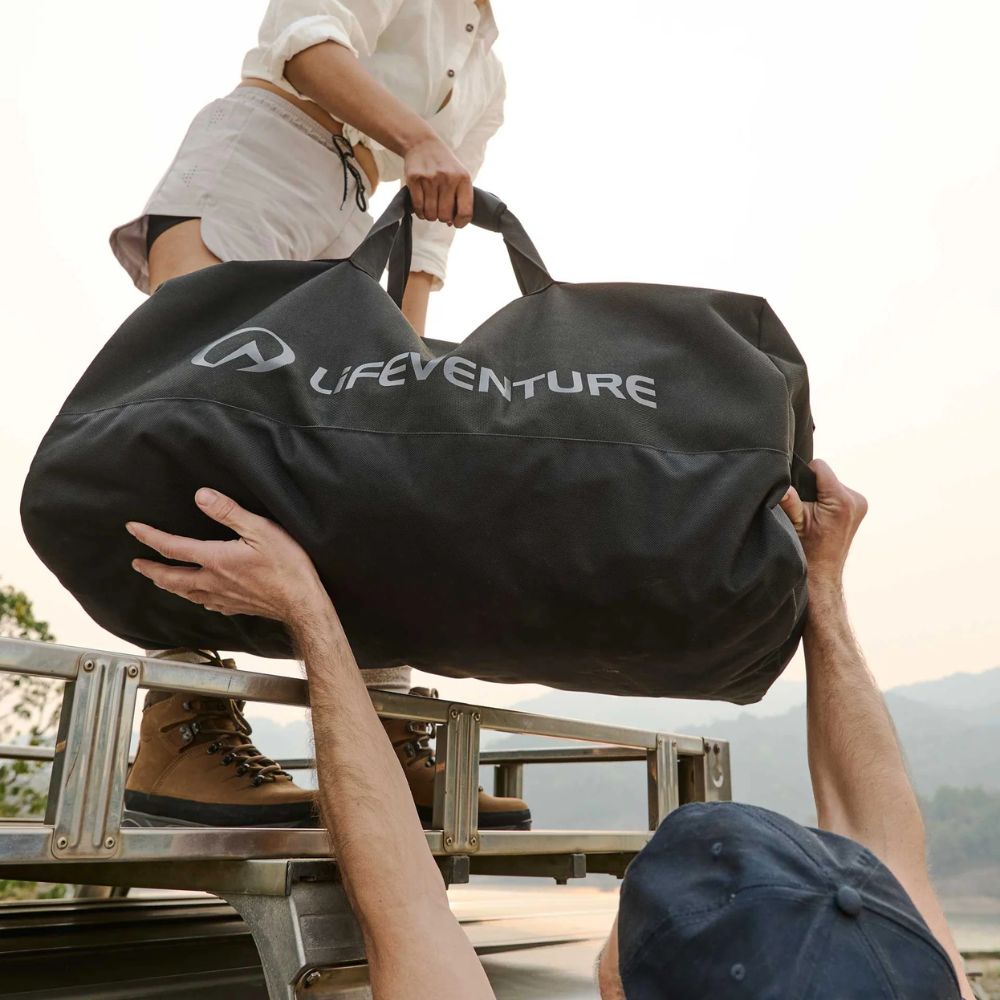 Two people lifting a black Lifeventure duffel bag onto a vehicle with a scenic background.