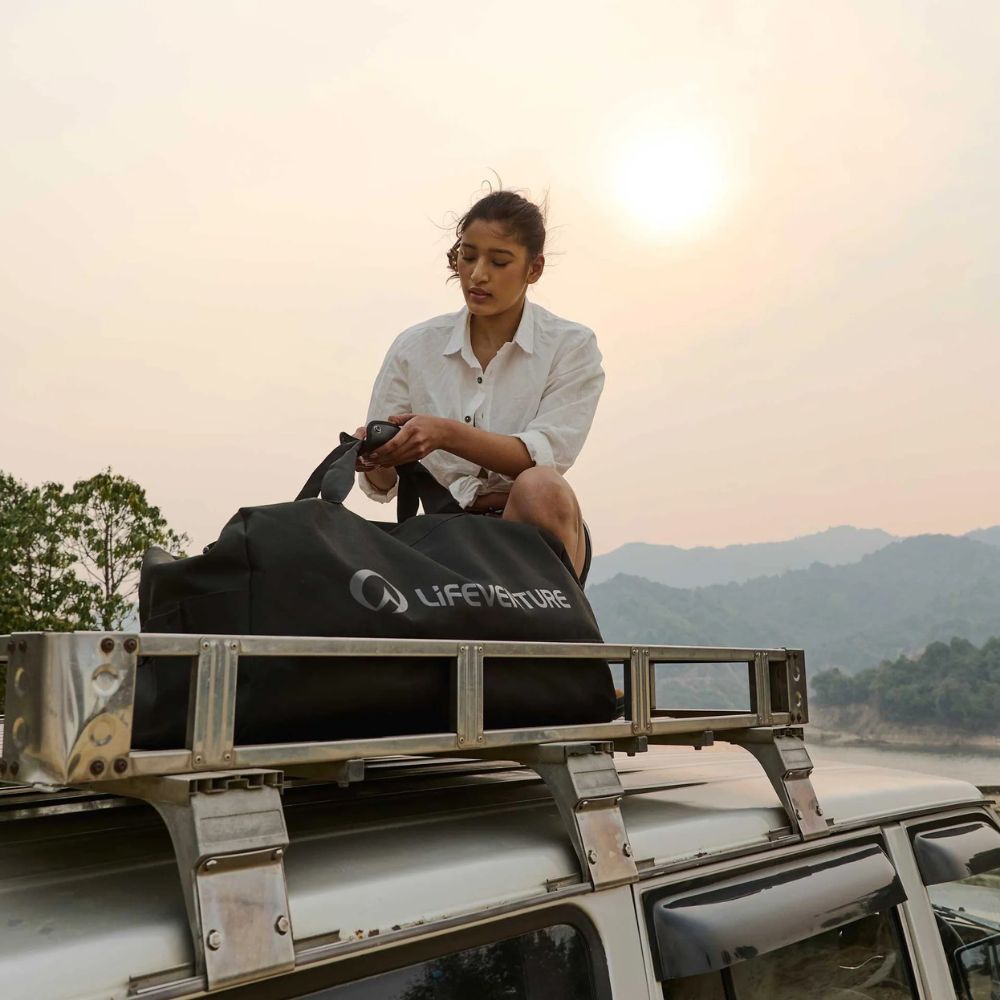 Person sitting on a vehicle with a Lifeventure bag, surrounded by nature.