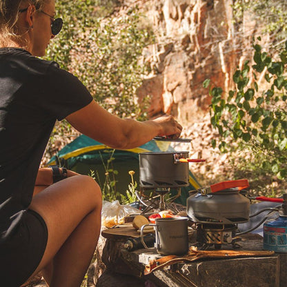 Person cooking outdoors using a portable stove with a scenic background