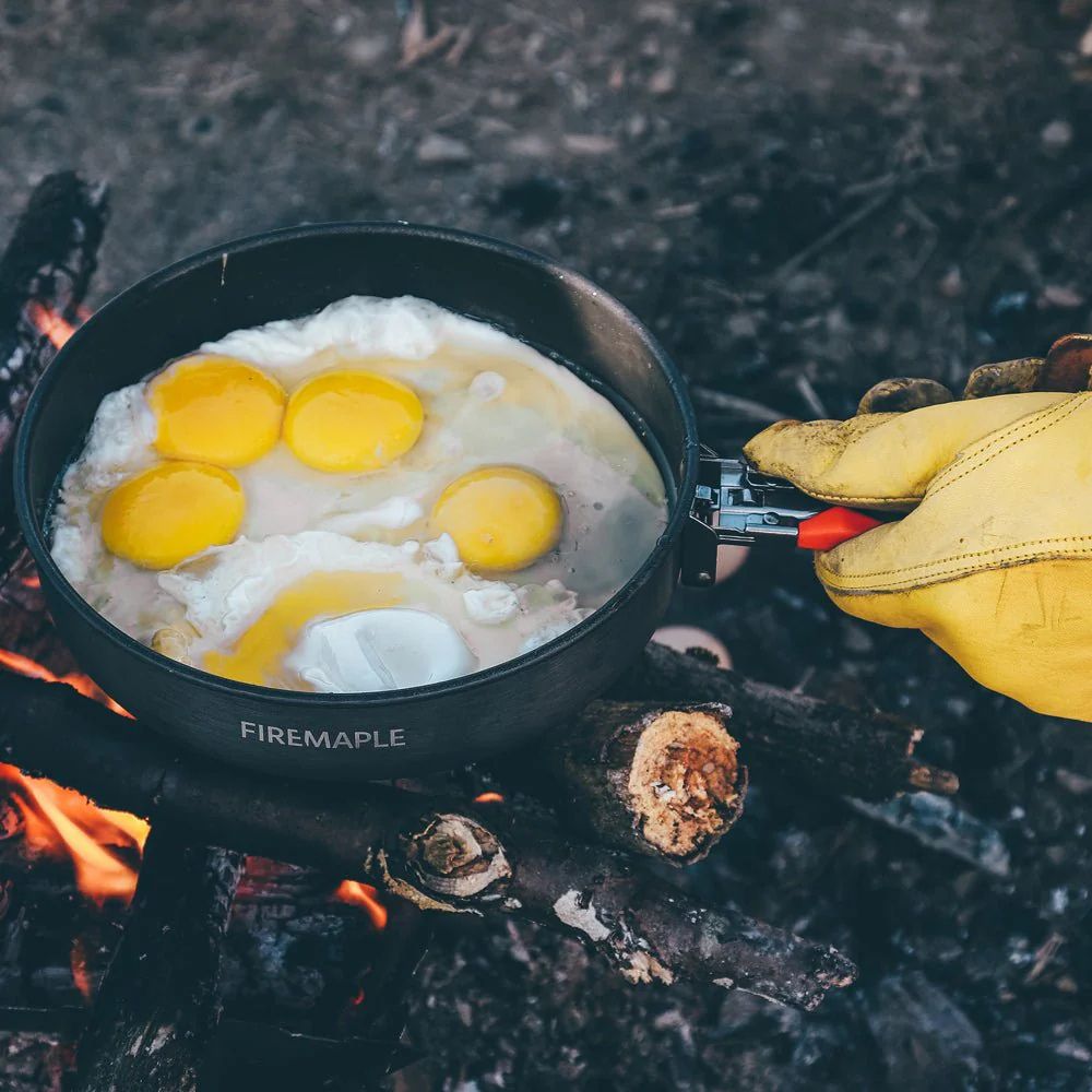 Frying pan with eggs over a campfire, held by a gloved hand.