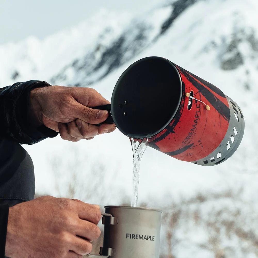 Person pouring liquid from a Firemaple camping stove into a mug with a snowy mountain background
