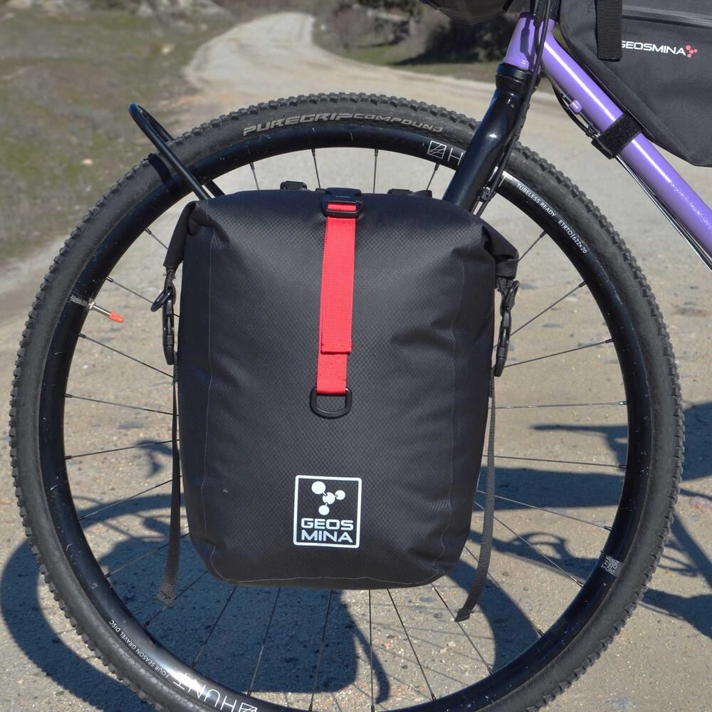 Black bike pannier with red strap attached to a bicycle wheel, featuring Geos Mina branding.