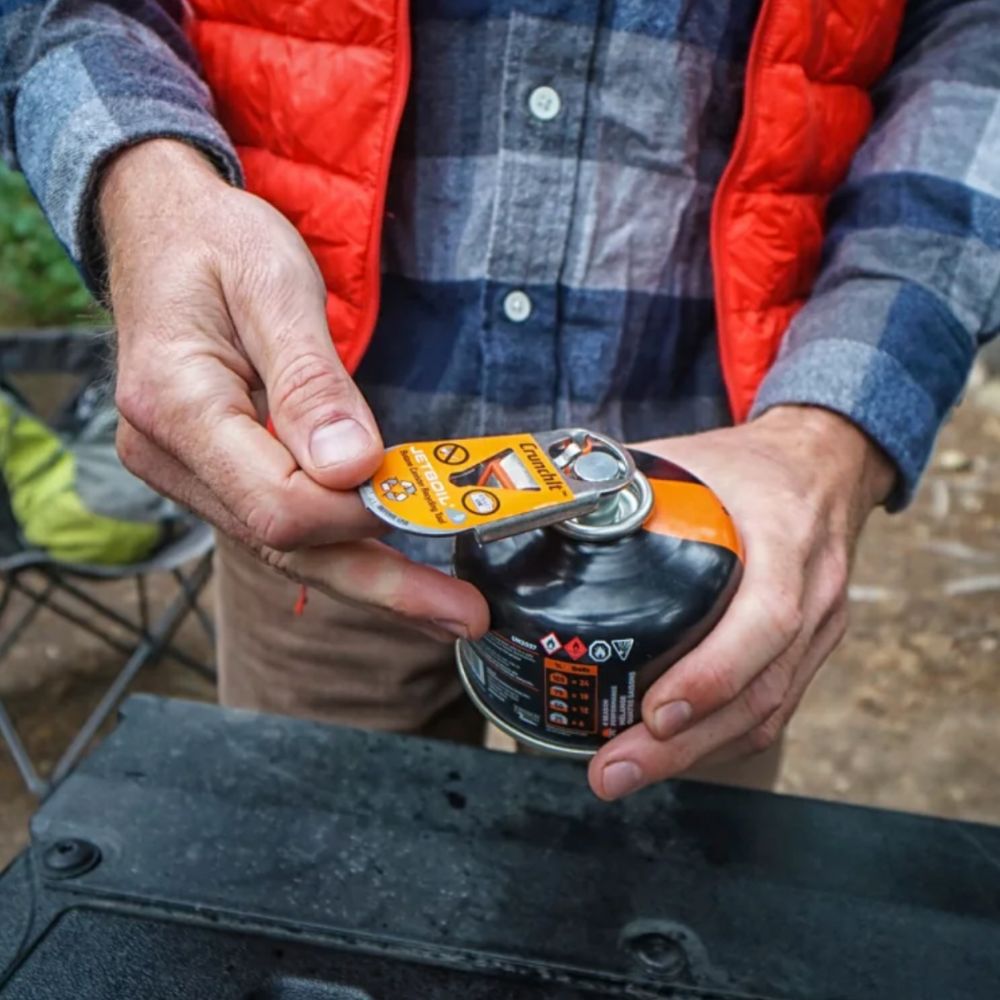 Person holding a camping stove with a red vest and plaid shirt in the background