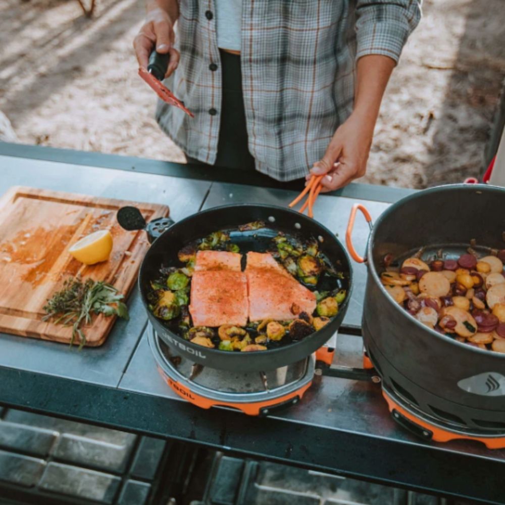 Person cooking outdoors with a portable stove and pan containing salmon and vegetables.