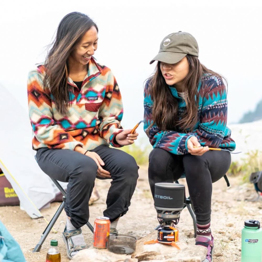 Two women sitting outdoors with camping gear, including a portable stove and drinks.