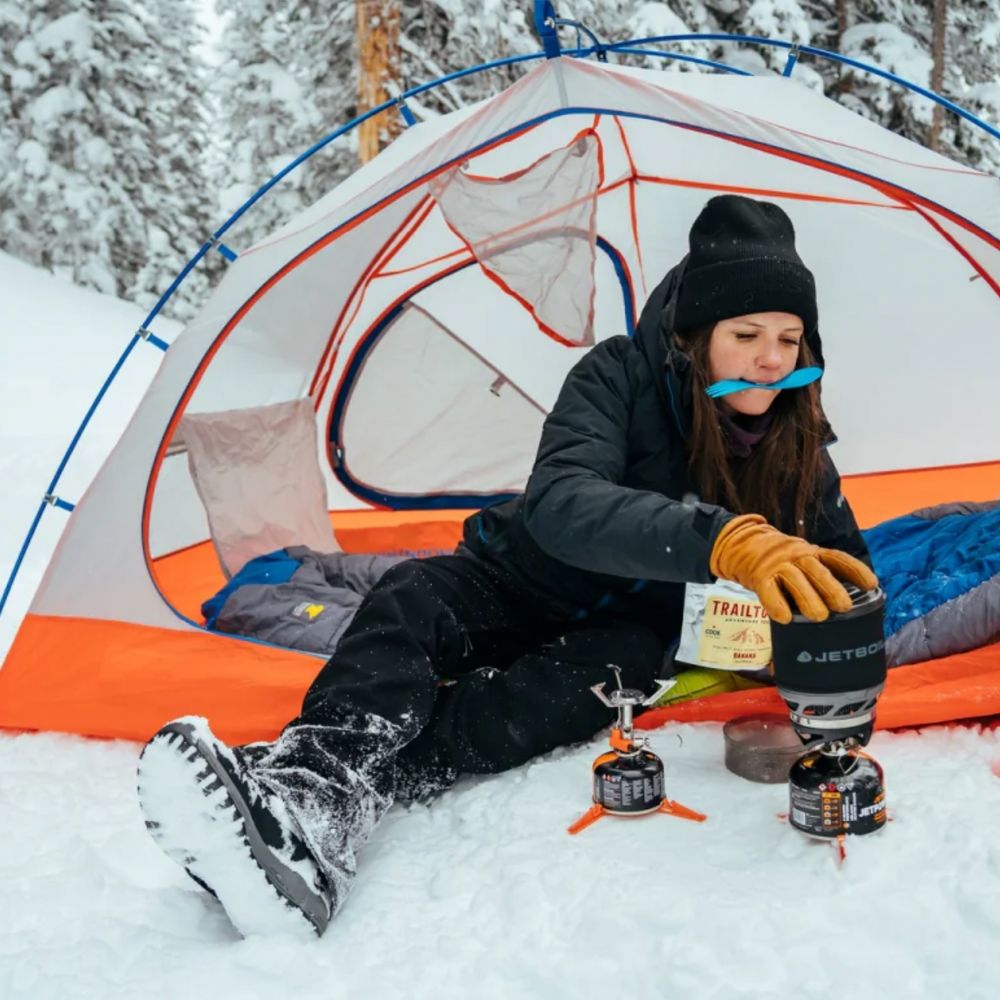 Person camping in the snow with a tent and camping stove.