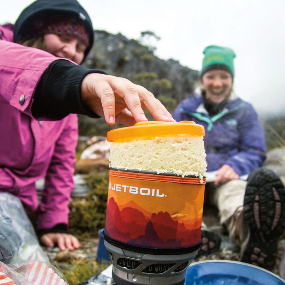 Person holding a JetBoil cooking system outdoors with two people in the background.