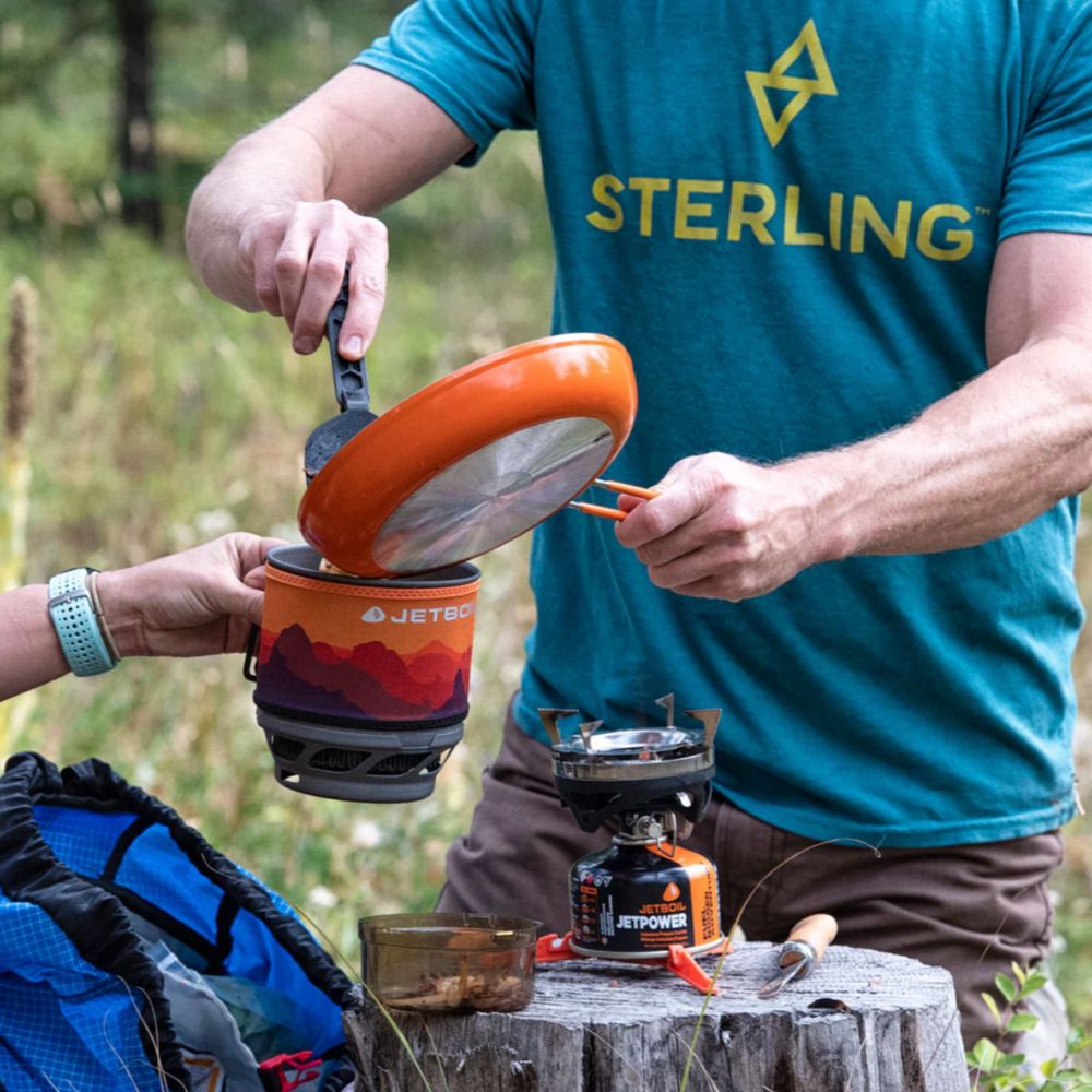 Person using a camping stove with a pot outdoors, wearing a blue 'Sterling' shirt.