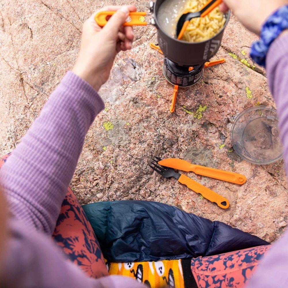 Person cooking outdoors using a portable stove with a pot on a rocky surface.