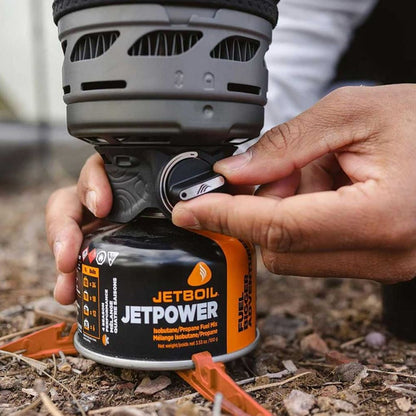 Person attaching a Jetboil fuel canister to a portable stove outdoors.
