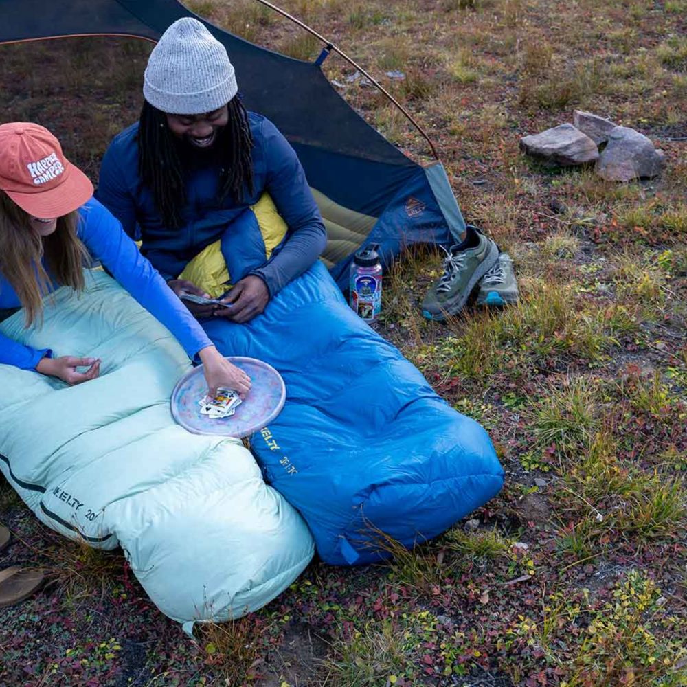Two people sitting in sleeping bags outdoors with a tent in the background