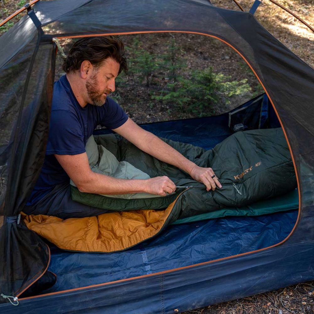 Man sitting inside a tent with a sleeping bag, surrounded by nature.