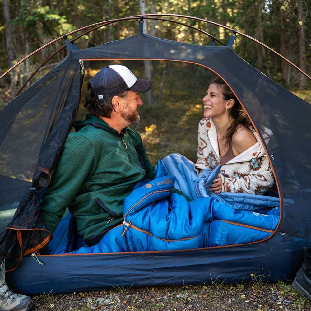 Two people sitting inside a tent with blue sleeping bags in a forest setting