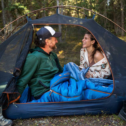 Two people sitting inside a tent with blue sleeping bags in a forest setting