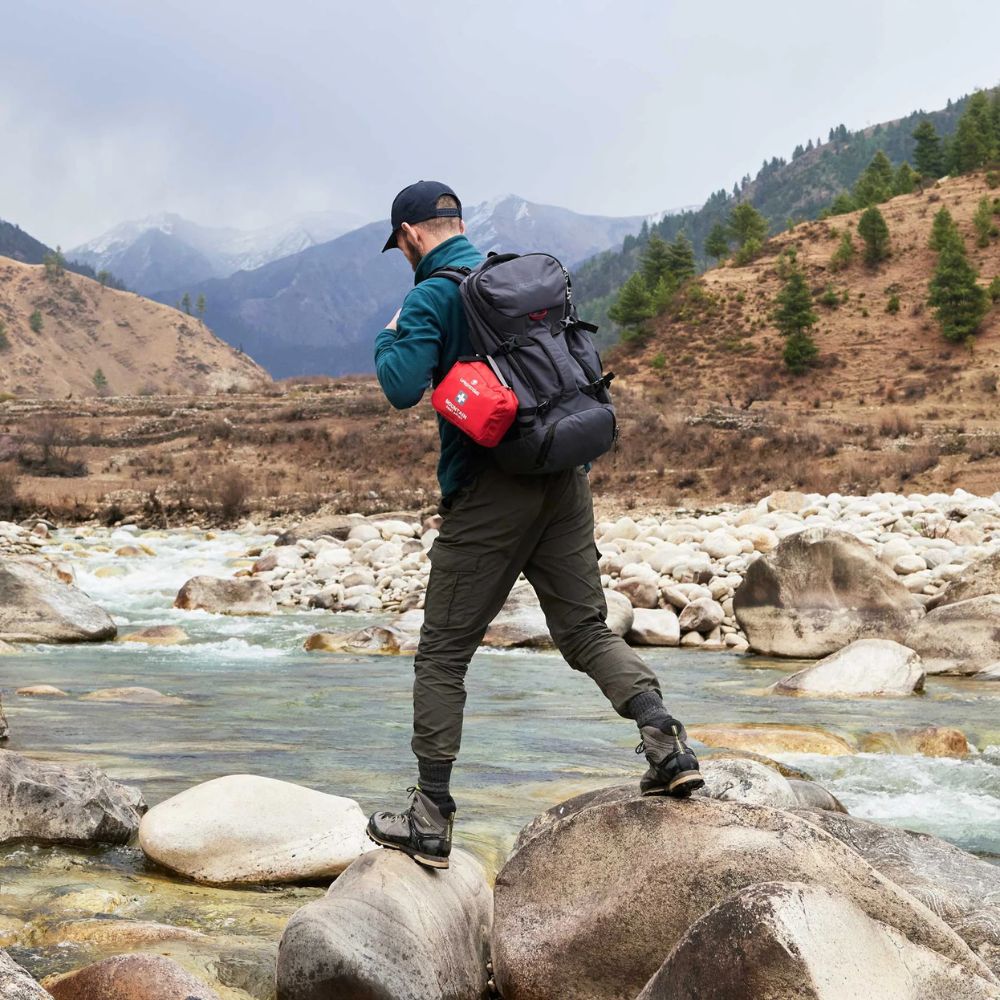 Person with a backpack standing on rocks by a river with mountains in the background