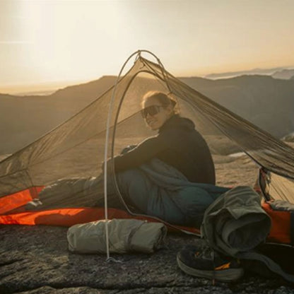 A woman wearing sunglasses sits inside a tent on a rocky mountain summit at sunset, with camping gear and a rolled sleeping bag in the foreground.