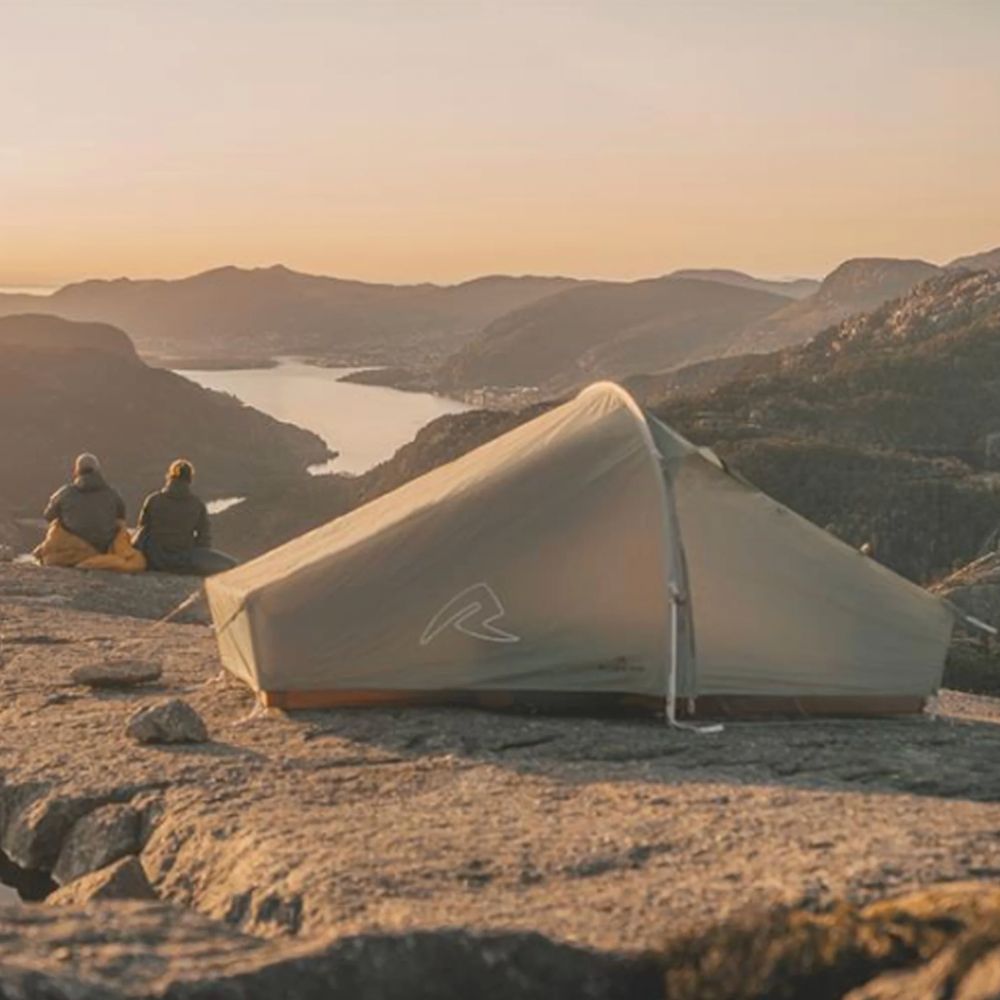 A large dome-style camping tent is pitched on a rocky mountain overlook at sunset, with two people sitting nearby and a fjord and mountains in the background.