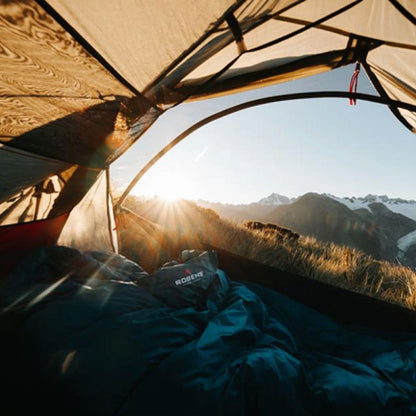 A camping sleeping bag and sleeping pad set inside a tent, with a mountain landscape visible outside under bright sun flare. A sleeping bag with the visible brand 'RODEWE' is placed in the foreground.