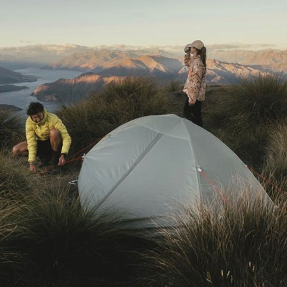 A light gray dome tent is pitched on a grassy mountain ridge with two people camping nearby, with a scenic lake and mountain range in the background at sunset.